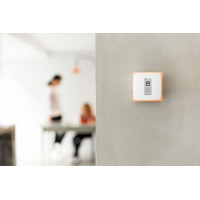 A digital thermostat displaying 19°C on a gray wall, with blurred figures of two people engaged in conversation nearby.