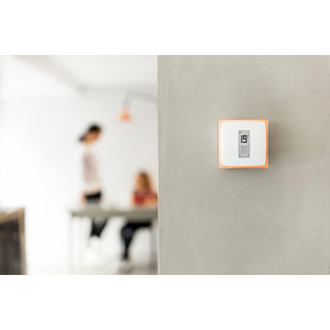 A digital thermostat displaying 19°C on a gray wall, with blurred figures of two people engaged in conversation nearby.